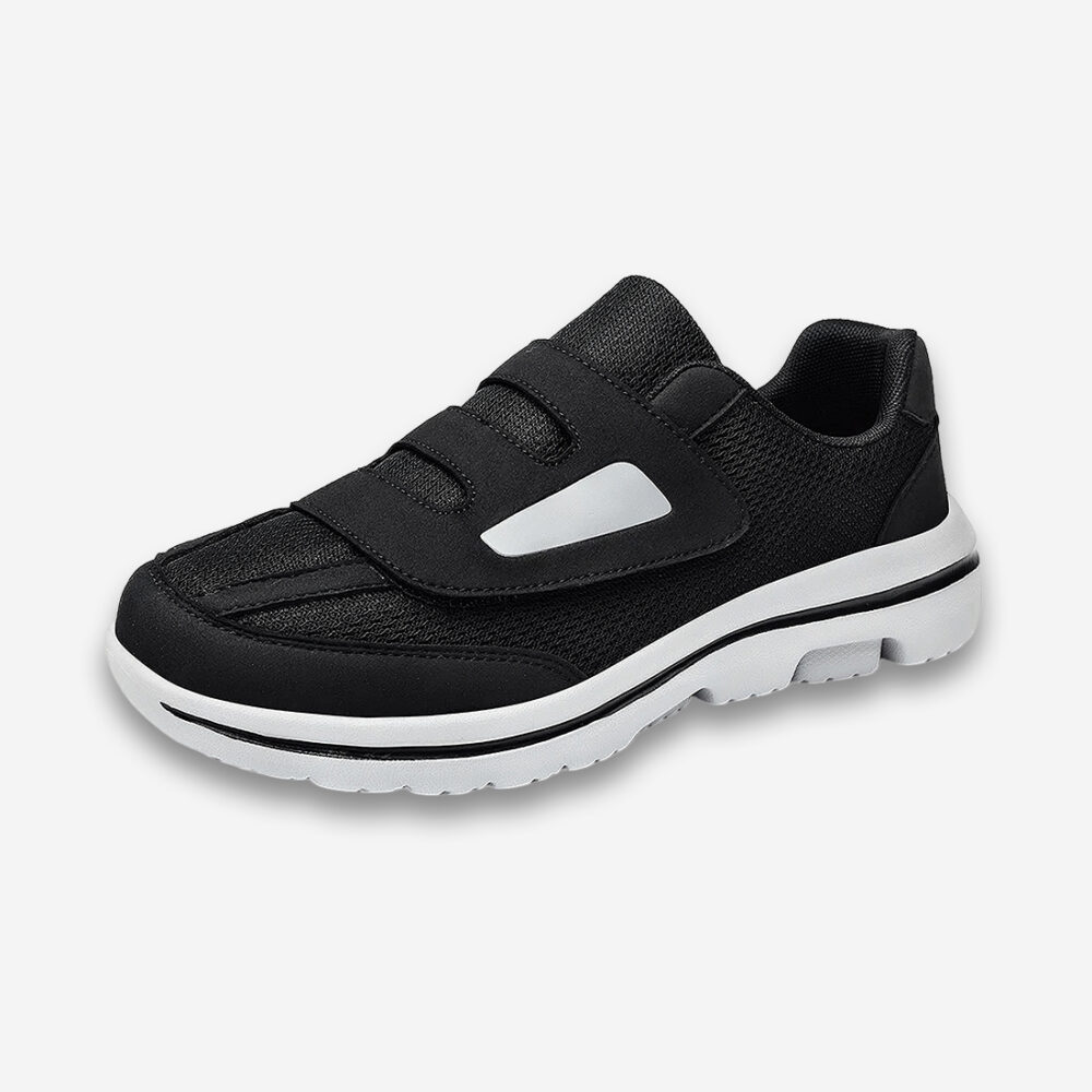 Une basket orthopédique homme facile à enfiler à velcro, de couleur noir et semelle blanche, disposée sur un fond gris très clair uni.