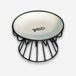 Bol orthopédique pour chat plat en céramique sur pied en métal noir façon grille. La coupelle en céramique est blanche avec un squelette de poisson au centre. Sur fond uni clair.