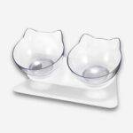 Gamelle orthopédique pour chat double bol incliné. Support blanc et deux bols en forme de tête de chat transparente. Sur fond uni clair.