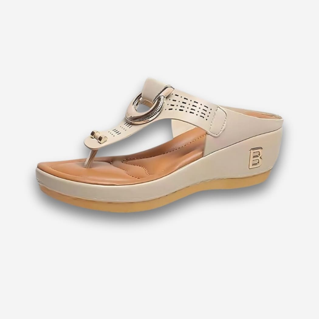 Sandales orthopédiques compensées pour femme beige avec détails dorés sur le dessus, ouvertes comme une tong avec le talon plus épais. Sur fond uni clair.