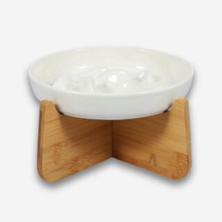 Bol orthopédique pour chat anti-glouton. La soucoupe est blanche en céramique avec des formes à l'intérieur pour séparer la nourriture. Elle est posée sur un support en bois clair. Sur fond uni clair.