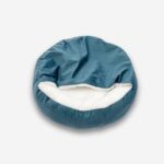 Lit orthopédique pour chien avec couverture intégrée. De forme ronde bleu turquoise avec l'intérieur blanc en peluche. Sur fond uni clair.