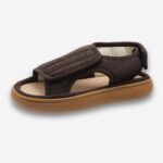 Chaussons orthopédiques large et ajustables pour femme de couleur marron avec une semelle en caoutchouc. Deux scratchs : un sur le dessus et un derrière. Le chausson est ouvert devant et sur les côtés. Sur fond gris très clair.