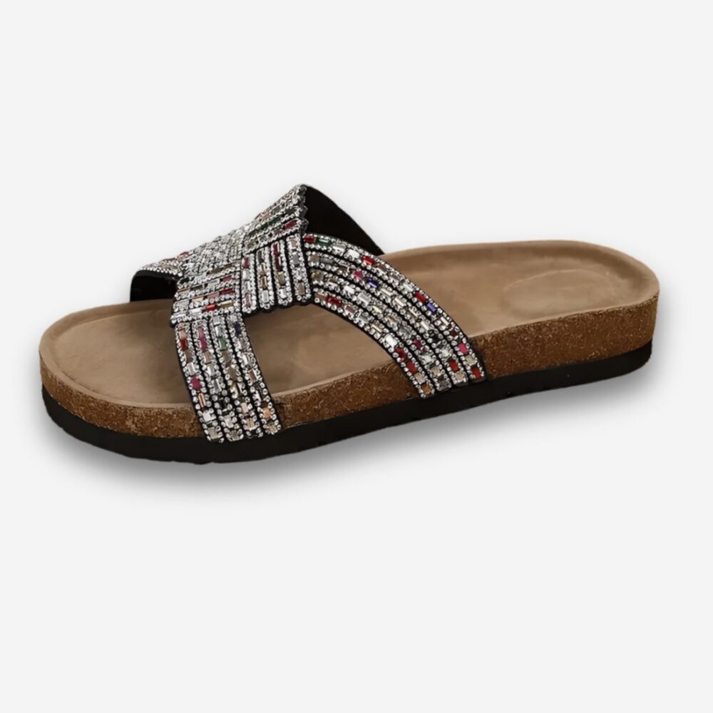 Mules orthopédiques pour femme à strass avec une semelle style en liège. Les brides croisées sont recouvertes de strass. Sur fond uni clair.
