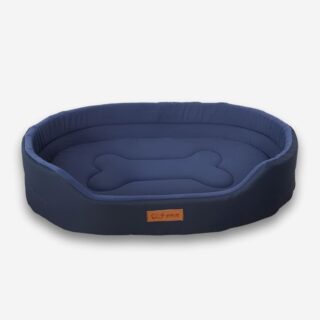 Panier orthopédique pour chien ovale et moelleux de couleur bleue avec un motif d'os au centre et une étiquette marron sur le devant. Sur fond uni clair.