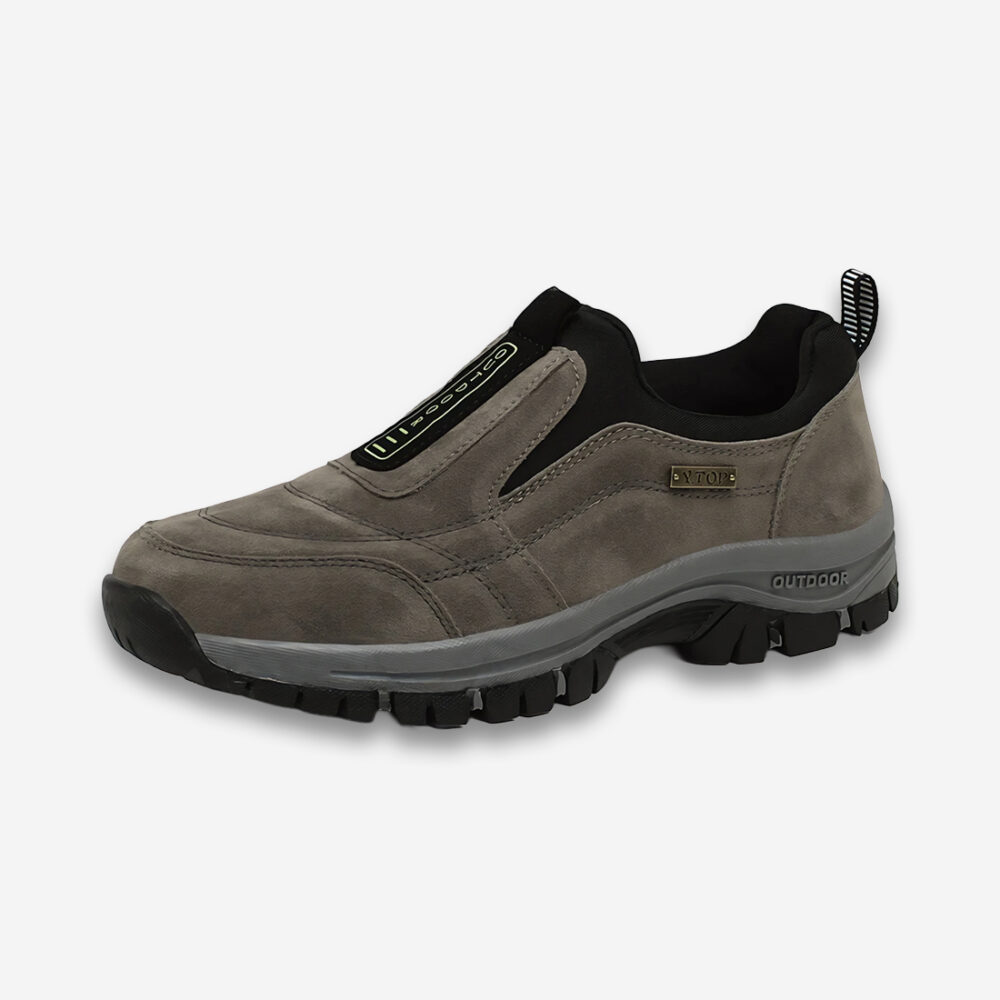 Une chaussure orthopédique homme de marche en daim, grise et noire, vue de profil sur un fond gris très clair uni.