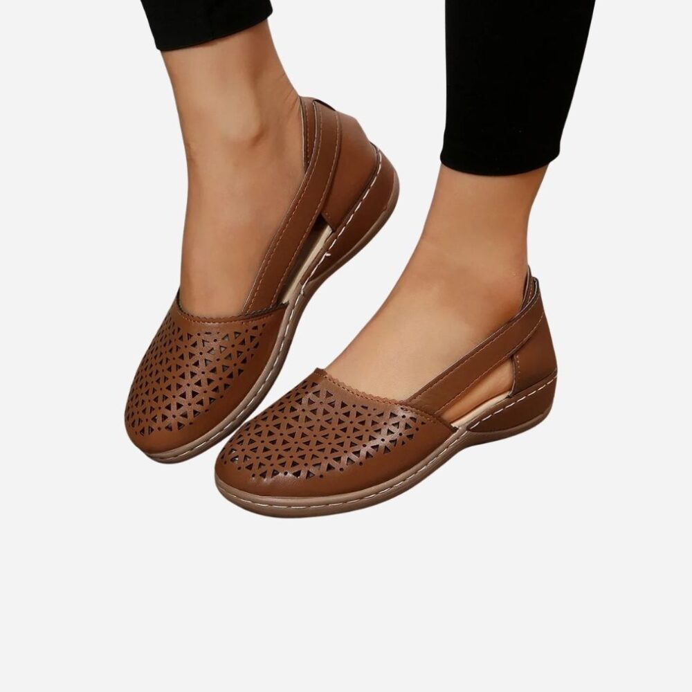 Chevilles d'une femme avec un pantalon noir et des sandales orthopédiques marrons aux pieds, avec des motifs ajourés et des semelles légèrement compensées.